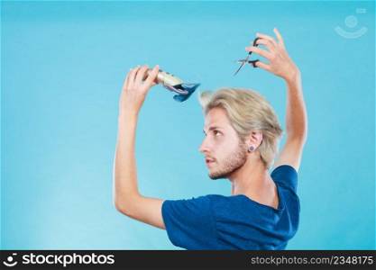 Hairdo changing hairstyle concept. Cool male hairstylist making self trendy haircut, fashion blonde metrosexual model going to shave his long hair, using shaver and scissors. Man going to shave his long hair