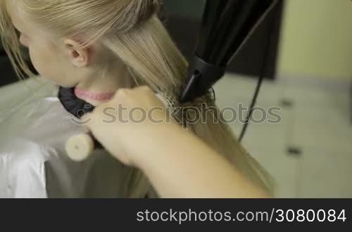 Hairdresser drying long blond straight hair of girl with hair dryer and round brush. Back view. Closeup. Hands of professional female hairstylist using round brush and blow dryer in barber shop.
