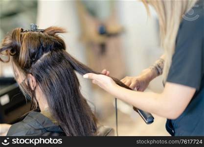 Hairdresser, protected by a mask, making waves in her client&rsquo;s hair with a hair iron in a salon. Business and beauty concepts. Hairdresser, protected by a mask, combing her client&rsquo;s hair with a hair iron in a salon.