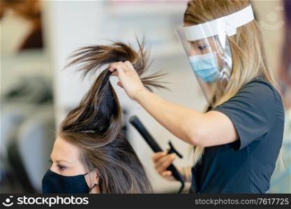 Hairdresser, protected by a mask, making waves in her client&rsquo;s hair with a hair iron in a salon. Business and beauty concepts. Hairdresser, protected by a mask, combing her client&rsquo;s hair with a hair iron in a salon.