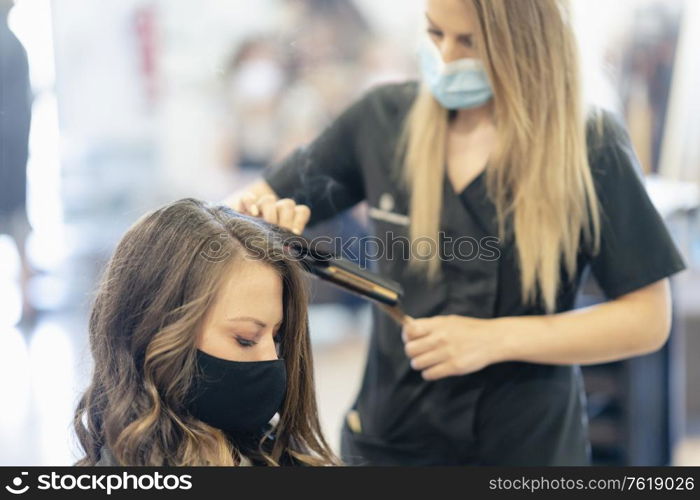 Hairdresser, protected by a mask, making waves in her client&rsquo;s hair with a hair iron in a salon. Business and beauty concepts. Hairdresser, protected by a mask, combing her client&rsquo;s hair with a hair iron in a salon.