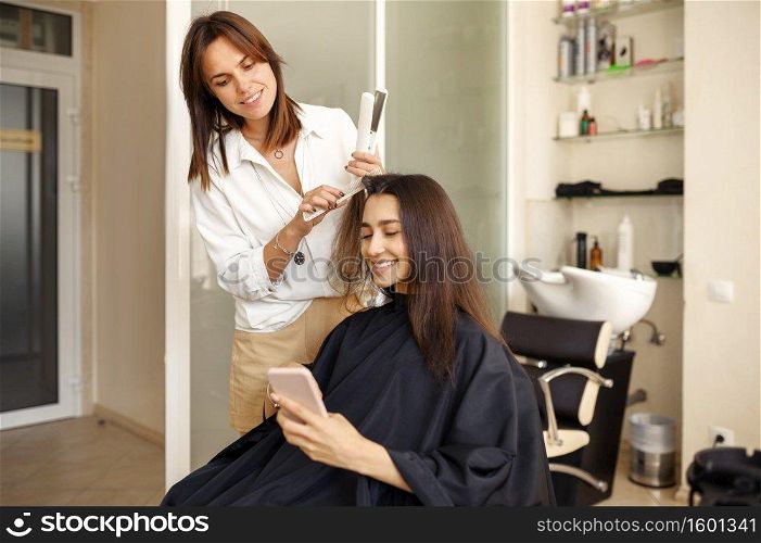 Hairdresser straightens woman’s hair, hairdressing salon. Stylist and client in hairsalon. Beauty business, professional service. Hairdresser straightens woman’s hair, hairsalon
