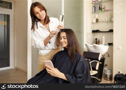 Hairdresser straightens woman’s hair, hairdressing salon. Stylist and client in hairsalon. Beauty business, professional service. Hairdresser straightens woman’s hair, hairsalon