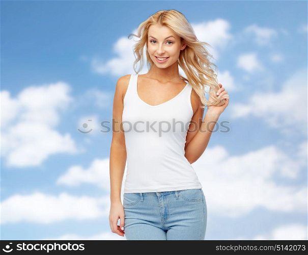 hairstyle, fashion and people concept - happy smiling beautiful young woman in white top and jeans with blonde hair over blue sky and clouds background. happy smiling young woman over blue sky