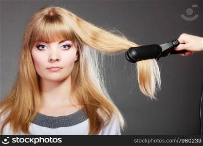 Hairstyling. Attractive blonde woman long haired making hairstyle hairdo with electric hair iron straightener gray background