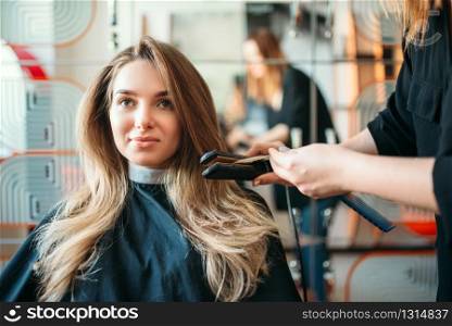 Hairstylist straightens hair with an iron, female client in hairdressing salon. Hairstyle making in beauty studio, hairdresser doing hairdo