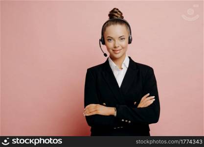 Half leghth shot of pretty female support center associate in dark suit wearing black wireless headset with mic being satisfied after solving customer problems, standing with arms crossed against pink wall. Happy female support center agent in dark suit posing in studio