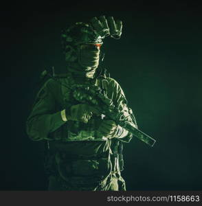 Half length, low key portrait of elite commando fighter, professional mercenary hiding identity behind mask, glasses, standing in darkness with mini submachine gun in hands, wearing nigh-vision device. Portrait of elite commando fighter in darkness