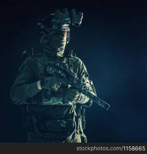 Half length, low key portrait of elite commando fighter, professional mercenary hiding identity behind mask, glasses, standing in darkness with mini submachine gun in hands, wearing nigh-vision device. Portrait of elite commando fighter in darkness