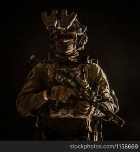 Half length, low key portrait of elite commando fighter, professional mercenary hiding identity behind mask, glasses, standing in darkness with mini submachine gun in hands, wearing nigh-vision device. Portrait of elite commando fighter in darkness