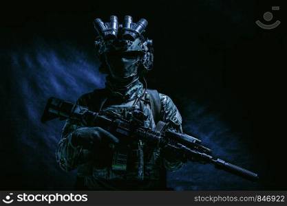 Half length, low key studio shoot of army soldier, marine infantryman in mask, camo uniform, equipped modern ammunition, armed service rifle standing in darkness with night vision device on helmet. Army special forces fighter low key studio shoot