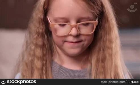 Half length of beautiful little girl in glasses with curly blonde long hair and blue eyes lifting up head, looking at camera and smiling. Happy and pretty facial expression.