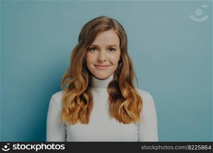 Half-length portrait of beautiful dreamy millennial woman with wavy hair in white turtleneck looking at camera with gentle smile isolated over pastel blue studio wall background with copy space. Beautiful dreamy young woman with perfect wavy styled hair in white turtleneck smiling at camera