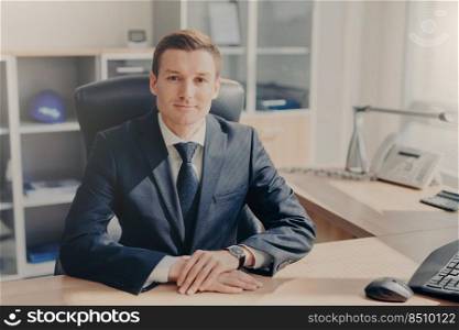 Half length portrait of handsome prosperous male manager sits at cabinet, looks directly into camera, has meeting with colleagues, discuss new business strategy. People and occupation concept