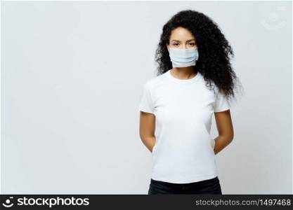 Half length shot of Afro American woman being on self isolation or quarantine, wears medical mask during coronavirus outbreak, listens news, tries not to spread virus, stays at home for long period