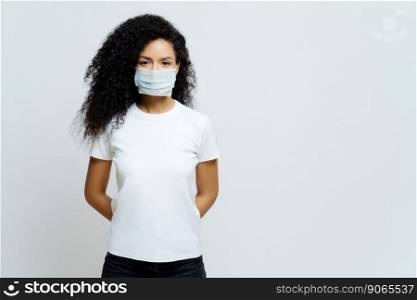 Half length shot of Afro American woman being on self isolation or quarantine, wears medical mask during coronavirus outbreak, listens news, tries not to spread virus, stays at home for long period