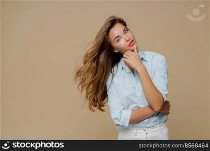 Half length shot of attractive female touches chin, keeps hands partly crossed over chest, tilts head, has long straight hair, dressed in stylish shirt and white jeans, poses against brown background