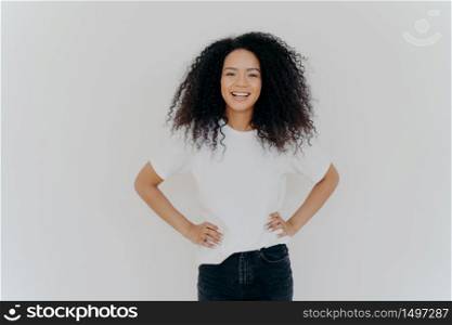 Half length shot of beautiful cheerful millennial girl stands with happy face expression against white studio background, keeps both hands on waist, has curly hair, minimal makeup, slim figure