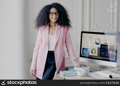 Half length shot of cheerful African American woman with curly hair, dressed in formal elegant outfit, poses near table, computer screen with graphics, prepares for presentation in front of auditorium