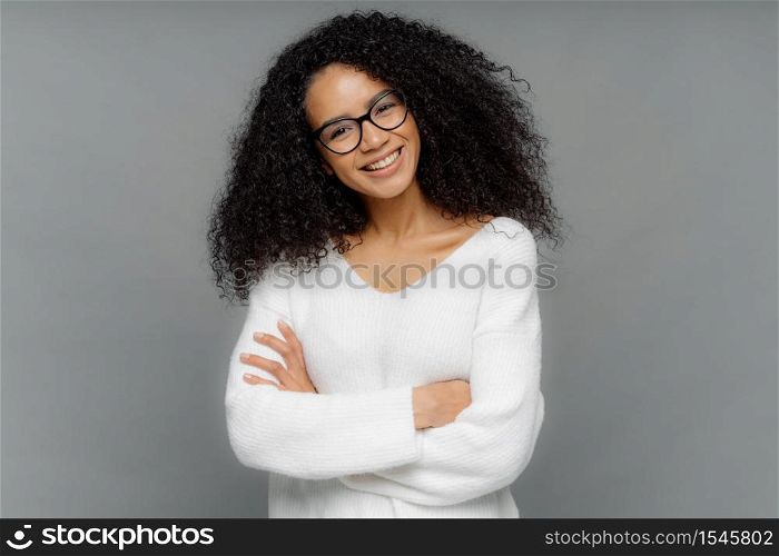 Half length shot of friendly looking African American woman tilts head, has dark crisp bushy hair, keeps arms folded over chest, wears casual white sweater, enjoys photoshoot in studio. Emotions