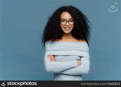 Half length shot of glad young female has confident facial expression, keeps arms folded, dressed in stylish sweater, happy to meet with colleagues, isolated on blue studio wall, blank space for text