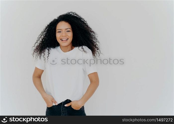Half length shot of good looking smiling woman laughs at funny joke, has fun, keeps both hands in pockets of jeans, wears white t shirt, has curly fluffy hair, poses indoor, blank space on right side