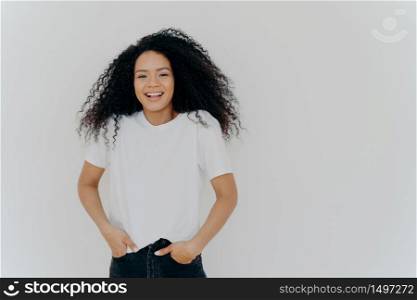 Half length shot of good looking smiling woman laughs at funny joke, has fun, keeps both hands in pockets of jeans, wears white t shirt, has curly fluffy hair, poses indoor, blank space on right side