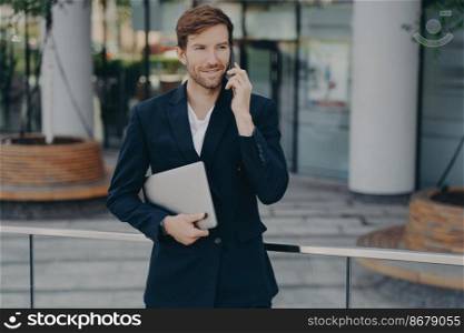Half length shot of handsome businessman makes phone call in roaming using tariffs carries digital device looks away into distance wears formal clothes. Executive manager has mobile talk outdoors. Executive manager inn formal clothes has mobile talk outdoors