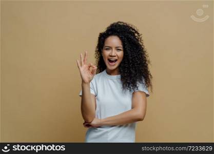 Half length shot of joyful curly young woman makes okay gesture, enjoys life and says ok, confirms everything is fine, wears white t shirt, isolated on beige background. Body language concept