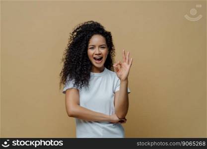 Half length shot of joyful curly young woman makes okay gesture, enjoys life and says ok, confirms everything is fine, wears white t shirt, isolated on beige background. Body language concept