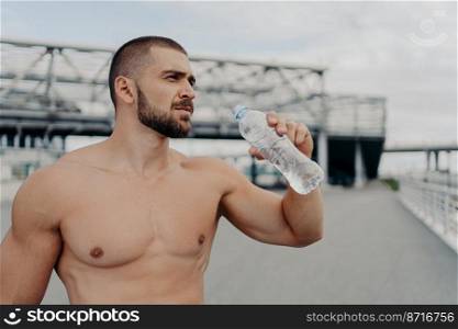 Half length shot of muscular guy drinks fresh water from bottle, takes rest after jogging exercising, has muscular arms, looks into distance thoughtfully, poses outside, cares about water balance