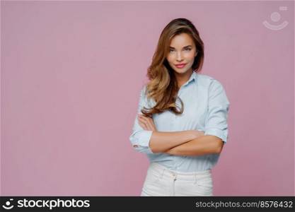 Half length shot of pleasant looking female model wears shirt and white jeans, keeps arms folded looks confidently at camera, has long hair, poses against purple background, blank space for promo