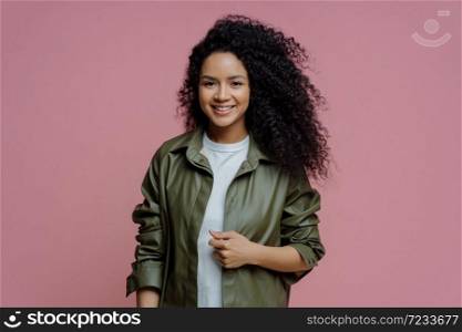 Half length shot of positive Afro American woman dressed in leather shirt, smiles pleasantly, enjoys good day, spends weekend with friends, has curly bushy hair, isolated on pink background.