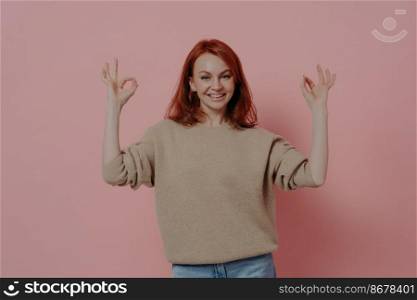 Half length shot of pretty ginger woman showing OK sign with both hands, saying that everything is fine, dressed in casual outfit, satisfied red-haired model posing isolated over pink background. Half length shot of pretty ginger woman showing OK sign with both hands, isolated on pink