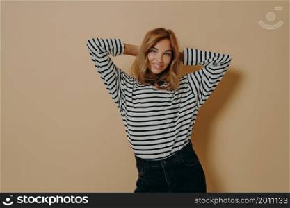 Half length shot of pretty satisfied young woman keeps hands behind head smiles gently wears striped jumper and black jeans isolated over beige background has natural beauty healthy clean skin. Half length shot of pretty satisfied young woman keeps hands behind head smiles gently poses indoor
