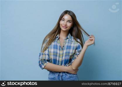 Half length shot of pretty young woman with dark hair, wears stylish clothes, has European appearance, applies makeup, poses in studio against blue background. People, beauty, lifestyle concept
