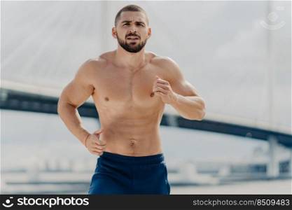 Half length shot of serious male jogger runs actively outdoor concentrated into distance poses with naked torso poses against blurred background with bridge. Fit muscular man runner trains actively
