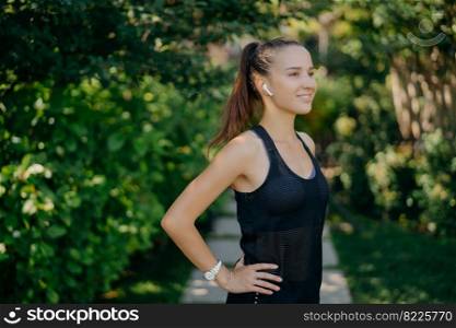 Half length shot of sporty young woman dressed in sportswear keeps hands on waist has dark hair combed in pony tail focused into distance with happy smile on face listens music poses outdoors