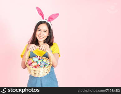 Half length studio shot of asian beauty happy young woman wearing bunny ears and holding colorful Easter egg in wood basket with lovely smile and colorful decor costume isolated on pastel backgrounds.