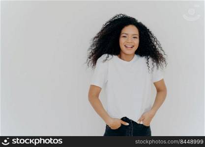Half≤n>h shot of good looking smiling woman laughs at funny joke, has fun, keeps both hands in pockets of jeans, wears white t shirt, has curly fluffy hair, poses indoor, blank space on right side