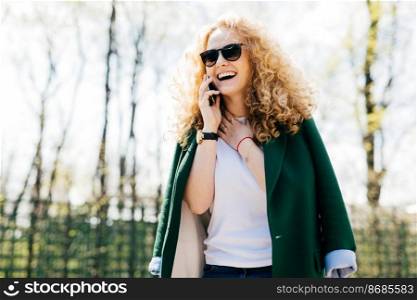 Half profile portrait of attractive woman with curly hair wearing stylish clothes and sunglasses talking on smart phone, smiling and laughing with happy and excited expression touching her neck