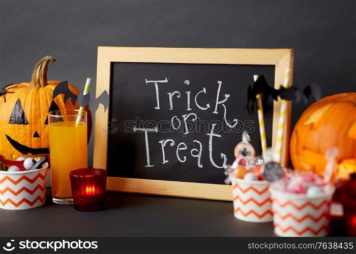halloween and holiday decorations concept - chalkboard with trick or treat lettering, jack-o-lanterns or carved pumpkins, candies, burning candles and glass of juice with paper straw. pumpkins, candies and halloween decorations