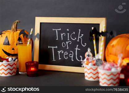 halloween and holiday decorations concept - chalkboard with trick or treat lettering, jack-o-lanterns or carved pumpkins, candies, burning candles and glass of juice with paper straw. pumpkins, candies and halloween decorations