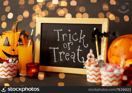 halloween and holiday decorations concept - chalkboard with trick or treat lettering, jack-o-lanterns or carved pumpkins, candies, burning candles and glass of juice with paper straw over bokeh lights. pumpkins, candies and halloween decorations
