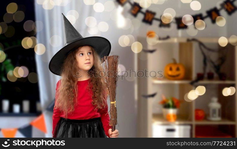 halloween, holiday and childhood concept - girl in costume and witch hat with broom over decorated home room and lights background. girl in black witch hat with broom on halloween
