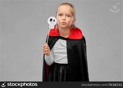 halloween, holiday and childhood concept - girl in dracula costume with black cape holding scull party accessory over grey background. girl in costume of dracula with cape on halloween