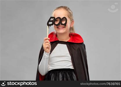 halloween, holiday and childhood concept - girl in dracula costume with black cape holding party accessory over grey background. girl in costume of dracula with cape on halloween