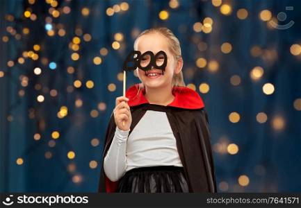halloween, holiday and childhood concept - girl in dracula costume with black cape holding party accessory over festive lights on dark blue background. girl in costume of dracula with cape on halloween