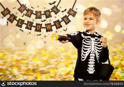 halloween, holiday and childhood concept - happy boy in black costume of skeleton with bucket of candies and flashlight trick-or-treating over garland string decoration and lights on background. happy with candies and flashlight on halloween