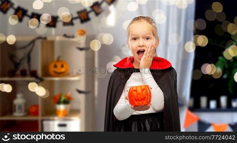 halloween, holiday and childhood concept - happy girl in black dracula cape or costume with jack-o-lantern pumpkin over decorated home room and lights background. girl in halloween costume of dracula with pumpkin